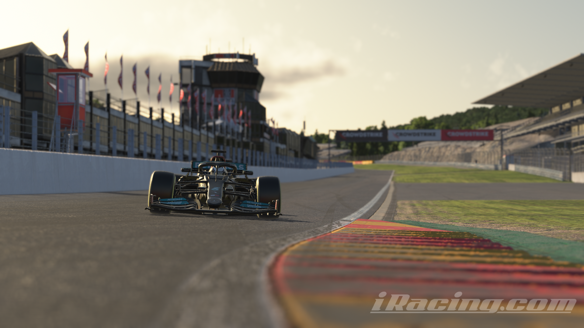 The Mercedes-AMG F1 W12 E Performance car approaches the famous Eau Rouge corner at Spa-Francorchamps during sunset. The car is positioned near the curb with brightly colored rumble strips on the right. In the background, pit buildings with flags and the iconic grandstands line the track, and the setting sun casts long shadows over the scene. iRacing screenshot.