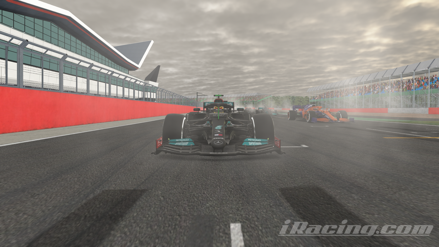 The Mercedes-AMG F1 W12 E Performance car races out of the starting grid position as lights go green at Circuit de Spa-Francorchamps on a cloudy day, with smoke from the tires trailing behind. The car is followed closely by a pack of other F1 cars, including an orange and blue car on the right. The grandstands are full of spectators and the sleek, red and white pit building is along the left side. iRacing screenshot.