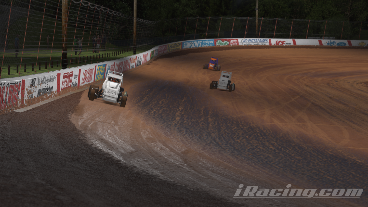 Several dirt sprint cars race around a banked oval dirt track at night. The number 5 white car leads the pack, sliding through the turn as dirt kicks up behind it. The track is lined with advertisements on the outer wall, and a small crowd of spectators watches from behind a fence under the dim lighting. iRacing screenshot.