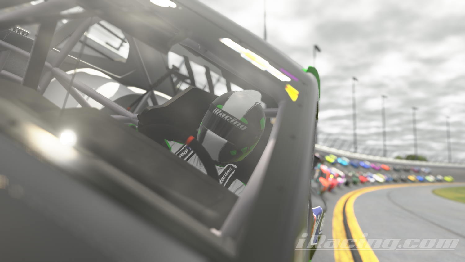 Close-up view of a NASCAR driver wearing a helmet with iRacing branding, seated inside the car and gripping the steering wheel. The camera focuses on the driver while the interior roll cage and race equipment are visible. In the background, a line of brightly colored Next Gen NASCAR Cup Series cars follow along during the pace lap on a banked corner of Daytona International Speedway under a cloudy sky. iRacing screenshot.
