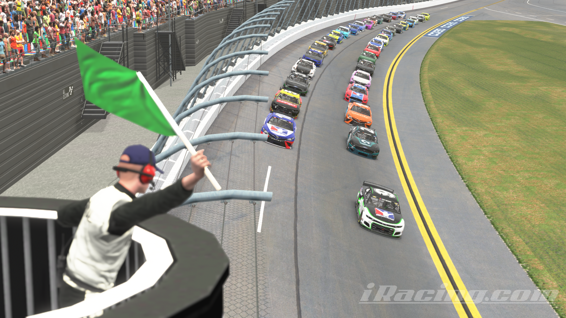 A field of Next Gen NASCAR Cup Series cars lines up for the start of a race at Daytona International Speedway. The green flag is being waved by an official in a raised flag stand, signaling the start of the race. The colorful cars are packed tightly together, and spectators fill the grandstands on the left, watching the action. The track is lined with safety barriers and guardrails, with grass on the infield. iRacing screenshot.