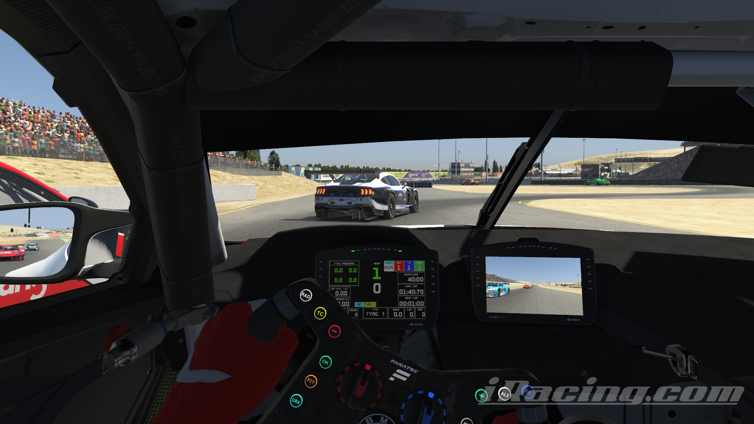Interior view from a race car cockpit during a road course race. The driver’s hands are visible on the steering wheel, with a digital dashboard displaying race information. A blue and white race car is directly ahead, while more cars can be seen through the side mirror and on the track. Spectators fill the stands on the hillside, and the race takes place on a sunny day with clear skies. iRacing screenshot.
