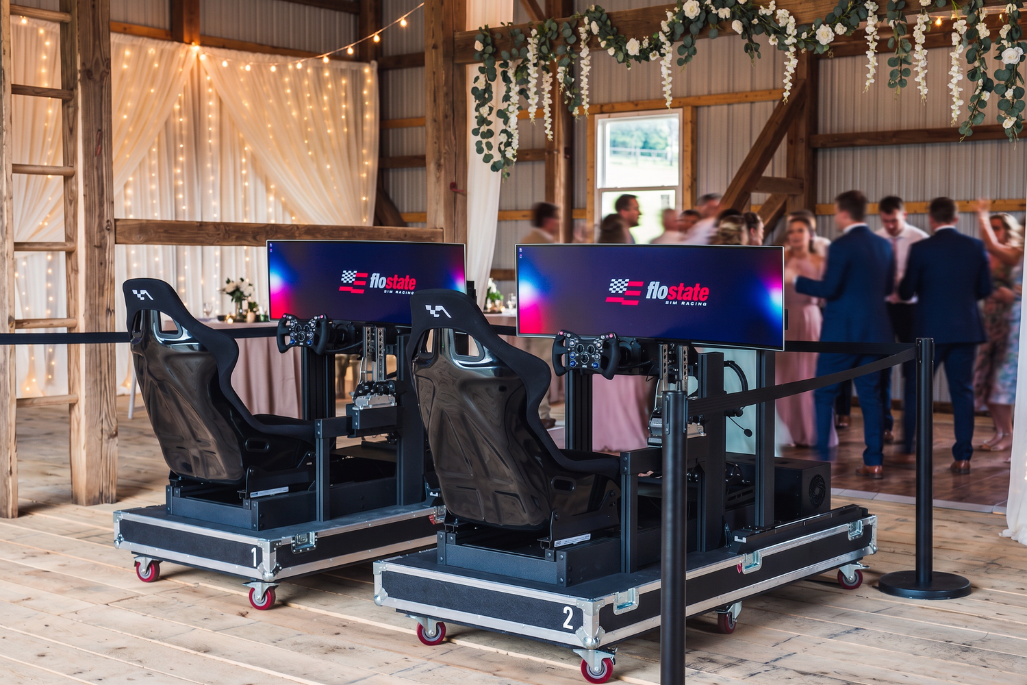Two racing simulator ride stations with black seats and screens, set up in a rustic barn event space with guests mingling in the background.